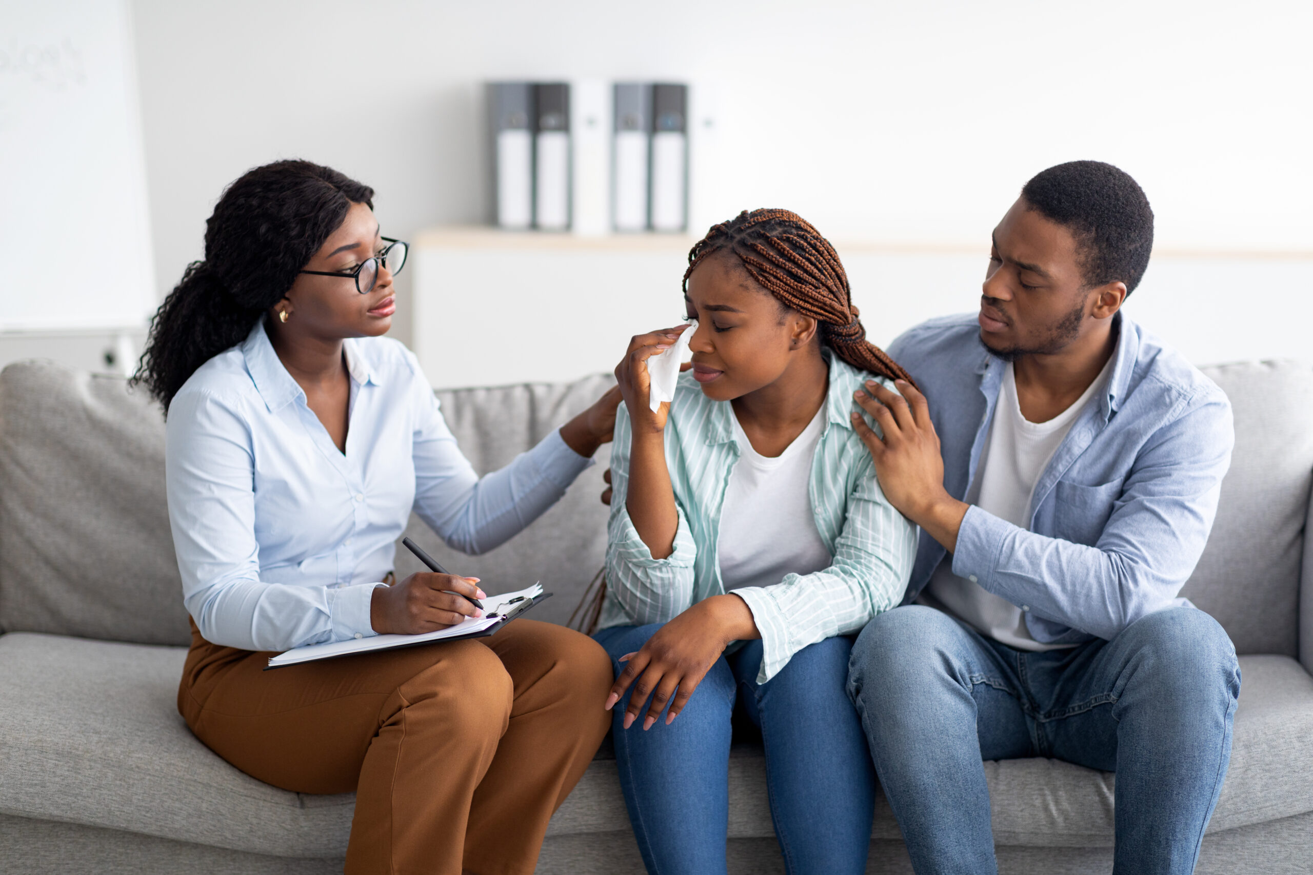 Millennial black woman crying at psychologist's office, loving husband supporting her with professional counsellor